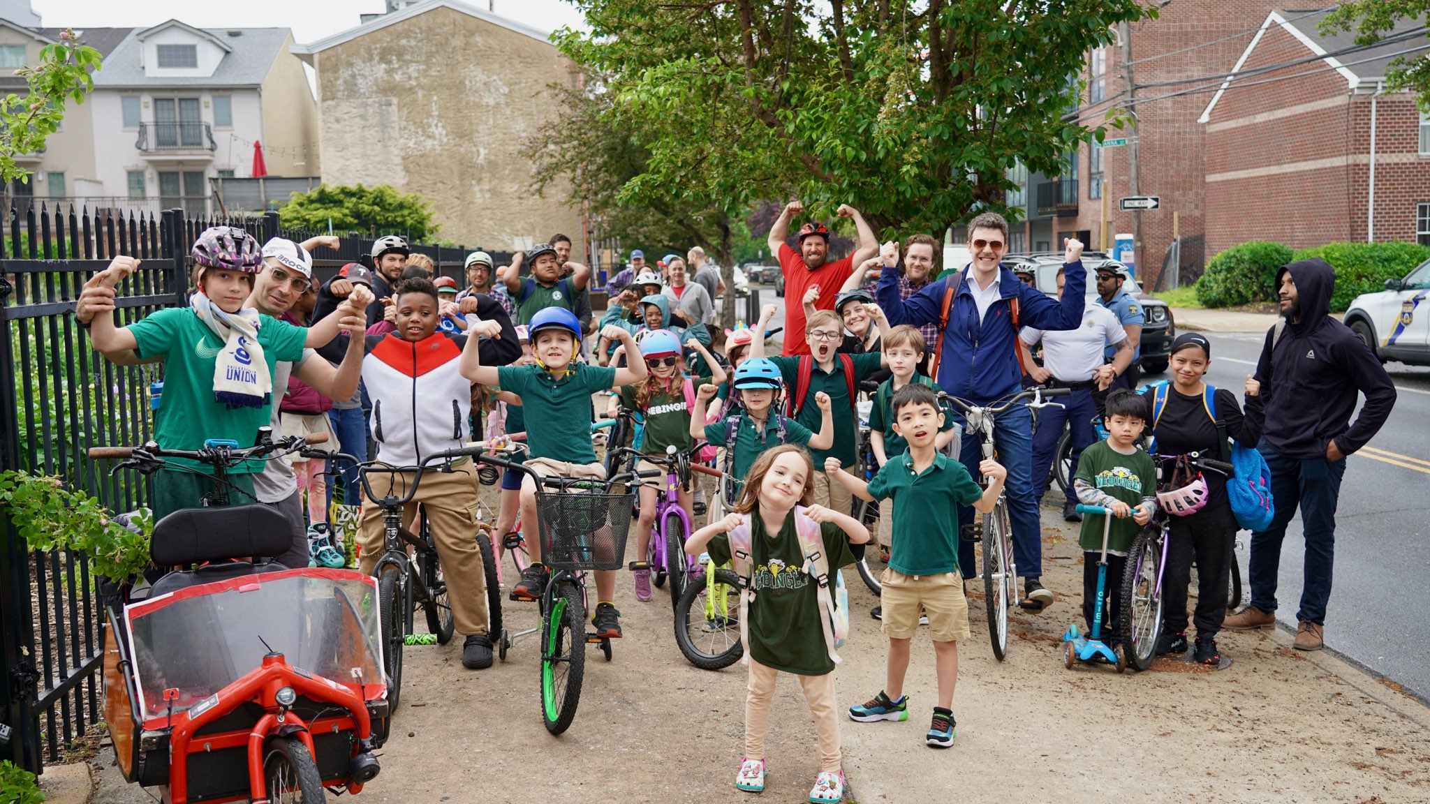 A group of children and adults pose with their bikes before riding to school. Many are holding up their arms in a flexing "strong" pose.